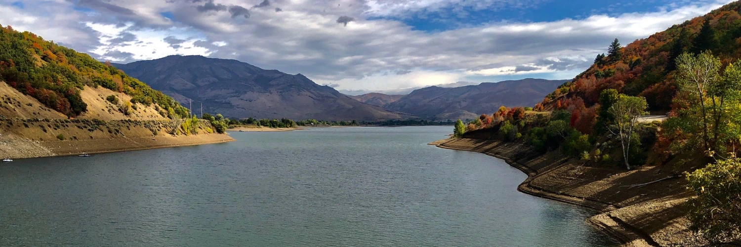 A calm reservoir lined with autumn trees, with mountains under a cloudy sky beyond it