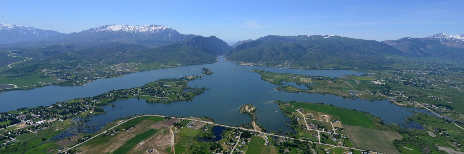 An aerial view of a large reservoir in a green mountain valley, with snow on the peaks behind it